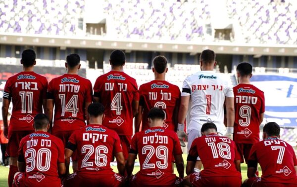 כל קבוצת הכדורגל הפועל תל אביב על המגרש עם הגב למצלמה ועם שמות הקיבוצים בעוטף ומספרים
